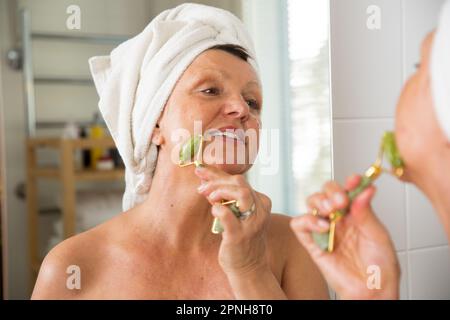 Mature belle femme faisant massage du visage avec rouleau de jade. Regarder dans le miroir dans la salle de bain, enveloppé dans une serviette. Routine soin de la peau et anti-âge Banque D'Images