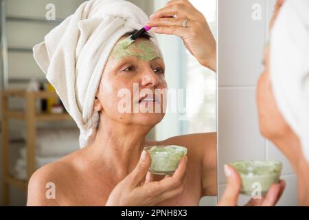 Mature belle femme appliquant le masque facial vert maison DIY sur la peau. Regarder dans le miroir dans la salle de bain, enveloppé dans une serviette. Routine de soin de la peau Banque D'Images
