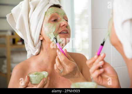 Mature belle femme appliquant le masque facial vert maison DIY sur la peau. Regarder dans le miroir dans la salle de bain, enveloppé dans une serviette. Routine de soin de la peau Banque D'Images