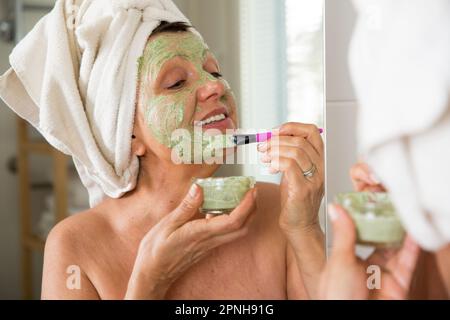 Mature belle femme appliquant le masque facial vert maison DIY sur la peau. Regarder dans le miroir dans la salle de bain, enveloppé dans une serviette. Routine de soin de la peau Banque D'Images