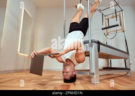Homme effectuant un exercice d'étirement de jambe unique pilates sur un lit de reformage dans une salle de gym en tournant pour faire face à la caméra pour étirer et tonifier les muscles abdominaux. Banque D'Images