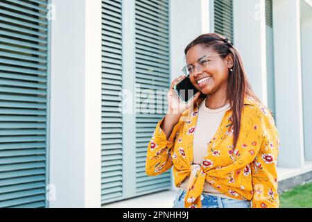 Une jeune étudiante afro-américaine parle sur un smartphone. Une femme hispanique ayant un appel de téléphone mobile debout à l'extérieur, riant et souriant dans une conversation amicale avec des camarades de classe d'université. Photo de haute qualité Banque D'Images