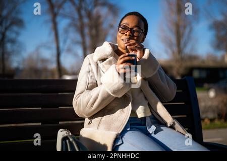 Portrait extérieur d'une femme noire heureuse le jour ensoleillé. Elle est assise sur un banc dans la rue et boit du café pour aller. Banque D'Images