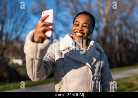 Portrait extérieur d'une femme noire heureuse le jour ensoleillé. Elle est assise sur le banc dans la rue et prend le selfie. Banque D'Images