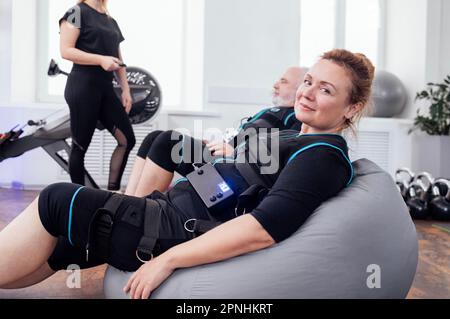 Un couple âgé souriant s'assoit sur des chaises de massage après une séance d'entraînement ems dans la salle de gym. Jeune femme entraîneur utilisant la télécommande définit les paramètres nécessaires sur electri Banque D'Images