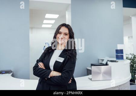 Une fille souriante de brune accueille à la réception dans un bureau moderne. La jeune femme en costume noir et en chemise blanche est debout près du comptoir semi-circulaire. PC portable, p Banque D'Images