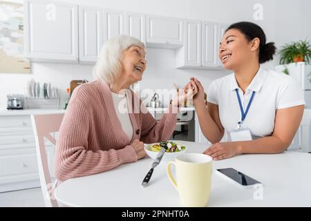 heureuse femme à la retraite avec cheveux gris tenant les mains avec joyeux travailleur social multiracial à côté de déjeuner sur table Banque D'Images