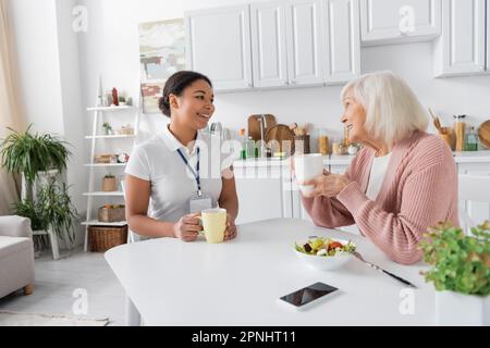 joyeux travailleur social multiracial ayant le thé avec une femme âgée dans la cuisine Banque D'Images