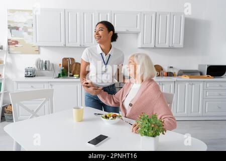 joyeux travailleur social multiracial tenant les mains d'une femme reconnaissante à la retraite dans la cuisine Banque D'Images