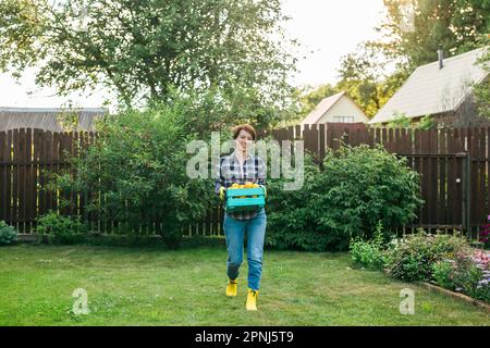 Fermier biologique tenant une boîte pleine de produits frais sur sa ferme. Une jeune femme heureuse souriant à l'appareil photo tout en se tenant dans son jardin potager. Réussite Banque D'Images