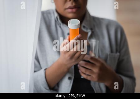 Vue rognée de la femme floue multiraciale tenant des pilules antidépresseurs et de l'eau près du rideau à la maison, image de stock Banque D'Images