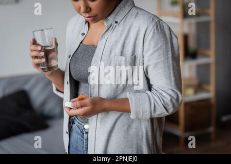 Vue rognée de la femme multiraciale tenant des pilules antidépresseurs et de l'eau à la maison, image de stock Banque D'Images