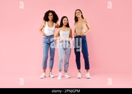 Trois jeunes femmes assez multiraciales debout et souriant à la caméra posant sur fond rose en studio, pleine longueur Banque D'Images