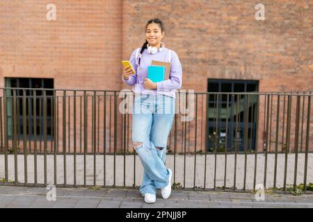 Fille d'université noire utilisant un smartphone et tenant des livres, debout à l'extérieur, pleine longueur Banque D'Images