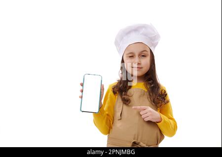 Petite fille portant le chapeau et le tablier du chef, pointe à blanc écran numérique de smartphone, en regardant l'appareil photo Banque D'Images