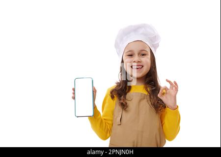 Une petite fille positive dans un chapeau de chef et un tablier, montrant un smartphone avec écran blanc vierge. Espace de copie pour les applications mobiles Banque D'Images