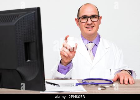 Un homme blanc casien chauve avec des lunettes portant un manteau blanc assis dans son bureau tenant un pot blanc de médecine. Banque D'Images