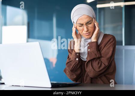 Fatigue au travail, surcharge. Une jeune femme arabe dans un hijab est assise dans le bureau à la table et tient sa tête, ressent de graves douleurs de tension, de pression, de massages avec ses mains. Banque D'Images