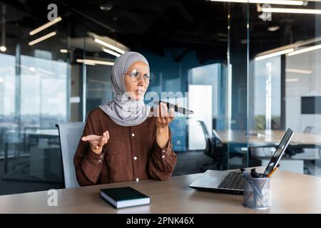 Inquiète jeune femme d'affaires musulmane dans le hijab assis au bureau et parlant au téléphone par haut-parleur, enregistrement de message vocal, résolution de questions, problèmes. Banque D'Images