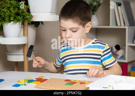 Un enfant d'âge préscolaire joue avec une mosaïque, construit des figures sur une planche, martelant des ongles avec un marteau sur des parties multicolores du concepteur Banque D'Images