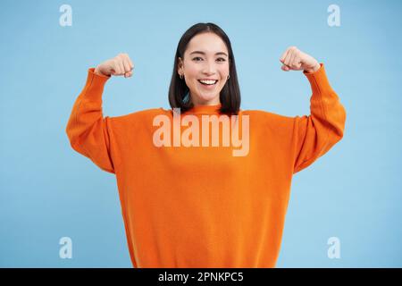 Des gens forts et en bonne santé. Une jeune femme asiatique montre ses muscles, fléchissant les biceps et semble confiant, souriant heureux, en grande forme, fond bleu Banque D'Images