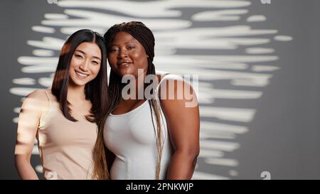 Des modèles de beauté souriants embrassant et regardant l'appareil photo, exprimant des émotions positives et des soins du corps. Belles jeunes femmes interraciales avec différents types de corps et skintones posant. Banque D'Images