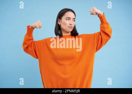 Portrait d'une femme coréenne mignonne en flexion de son biceps, montre des bras forts, des muscles et des sourires, concept de personnes en bonne santé et en forme, fond bleu Banque D'Images