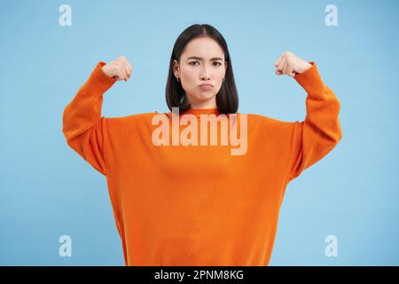 Des gens forts et en bonne santé. Une jeune femme asiatique montre ses muscles, fléchissant les biceps et semble confiant, souriant heureux, en grande forme, fond bleu Banque D'Images