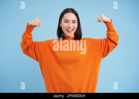 Des gens forts et en bonne santé. Une jeune femme asiatique montre ses muscles, fléchissant les biceps et semble confiant, souriant heureux, en grande forme, fond bleu Banque D'Images