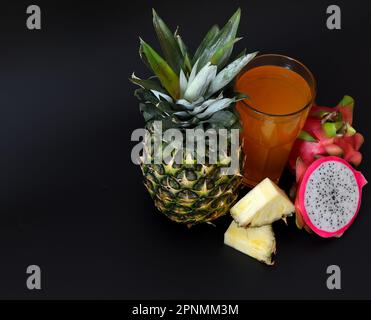 Un grand verre de jus de fruits mélangés sur fond noir, à côté d'un ananas mûr et de morceaux de pitaya. Gros plan. Banque D'Images