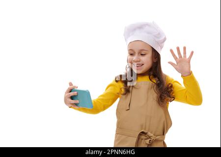 Sourire positif petite fille dans le chapeau de chef blanc et tablier de cuisine beige agitant avec la main pendant la vidéo-conférence Banque D'Images