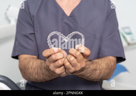 Rognez un dentiste masculin non reconnaissable en uniforme en faisant la forme du cœur avec des dispositifs de retenue en silicone transparent dans une clinique moderne Banque D'Images