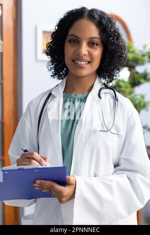 Portrait d'une femme biraciale heureuse avec un presse-papiers et souriant au bureau du médecin Banque D'Images