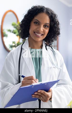 Portrait d'une femme biraciale heureuse avec un presse-papiers et souriant au bureau du médecin Banque D'Images
