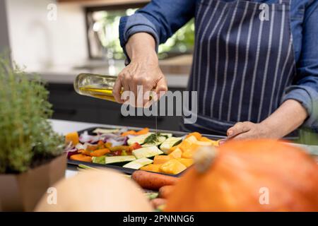 Section médiane de la femme caucasienne âgée versant de l'huile d'olive sur des légumes dans la cuisine Banque D'Images