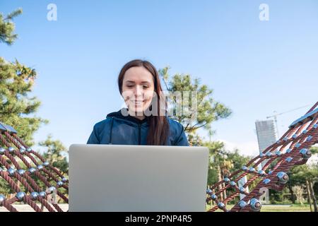 Jeune femme caucasienne, nomade numérique, indépendante travaillant à distance en plein air assis Oh hamac, en utilisant un ordinateur portable, souriant. Chat féminin joyeux au zoom ou Banque D'Images