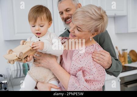 bébé fille jouant avec la voiture de jouet près de grands-parents heureux dans la cuisine, image de stock Banque D'Images