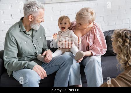 enfant sans souci riant près de grands-parents heureux et mère floue dans le salon, image de stock Banque D'Images