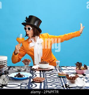 Femme impressionnante et excentrique dans des vêtements élégants, cylindre et lunettes de soleil, boire du jus sur la table pleine de chocolat. Banque D'Images