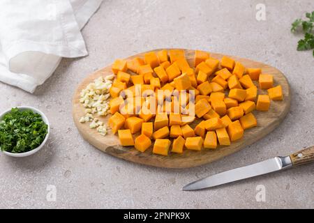 Planche en bois avec cubes de citrouille et ail haché avec persil sur fond beige texturé, vue du dessus. Cuisine délicieuse nourriture végétalienne Banque D'Images