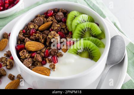Yaourt grec avec granola au chocolat, kiwi, graines de grenade et amandes dans un bol pour un petit déjeuner sain sur fond blanc. Gros plan Banque D'Images