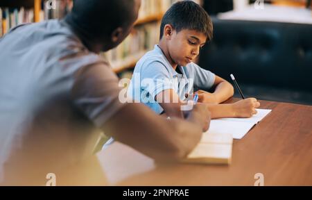Services de counselling aux enfants à l'école primaire. Un jeune garçon d'école écrit dans un journal tout en étant assis dans une bibliothèque avec un éducateur. Élève de l'école primaire havi Banque D'Images