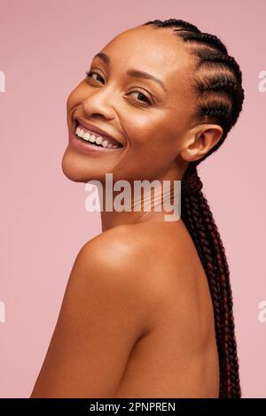 Modèle féminin attrayant avec une peau brillante, qui regarde l'appareil photo et sourit. Femme afro-américaine avec une peau impeccable sur fond rose. Banque D'Images