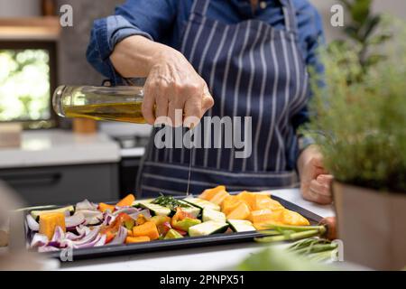 Section médiane de la femme caucasienne âgée versant de l'huile d'olive sur des légumes dans la cuisine Banque D'Images