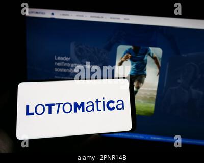 Personne tenant le téléphone portable avec le logo de la société italienne de jeu Lottomatica spa à l'écran en face de la page web d'affaires. Mise au point sur l'affichage du téléphone. Banque D'Images