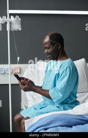 Image verticale de l'homme afro-américain avec compte-gouttes communiquant en ligne à l'aide de son smartphone tout en étant assis sur le lit dans le quartier Banque D'Images