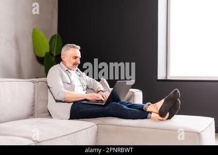 Beau homme d'âge moyen assis à l'aide d'un ordinateur portable avec un sourire heureux pendant qu'il se détend sur un canapé confortable à la maison Banque D'Images