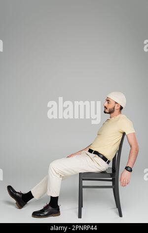 Vue latérale de l'homme gay barbu en chapeau beige assis sur une chaise sur fond gris, image de stock Banque D'Images
