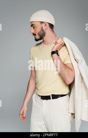 Vue latérale de l'élégant homme gay dans le chapeau tenant la veste et regardant loin isolé sur gris, image de stock Banque D'Images