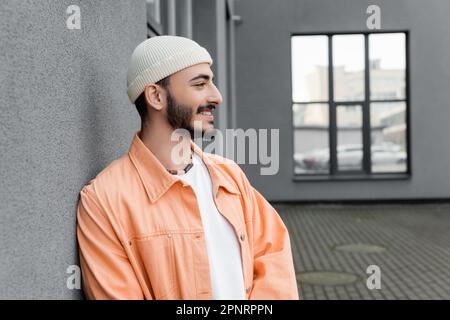 Vue latérale de l'homme gay insouciant dans le chapeau regardant loin de bâtiment sur la rue urbaine, image de stock Banque D'Images